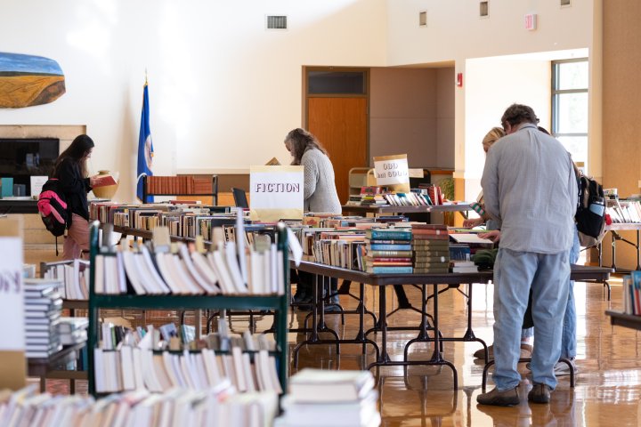 People browsing through tables filled with books at a book sale. Signs label different sections, including 'Fiction' and 'Odd but Good.' The room is brightly lit, and a Minnesota state flag is visible in the background.